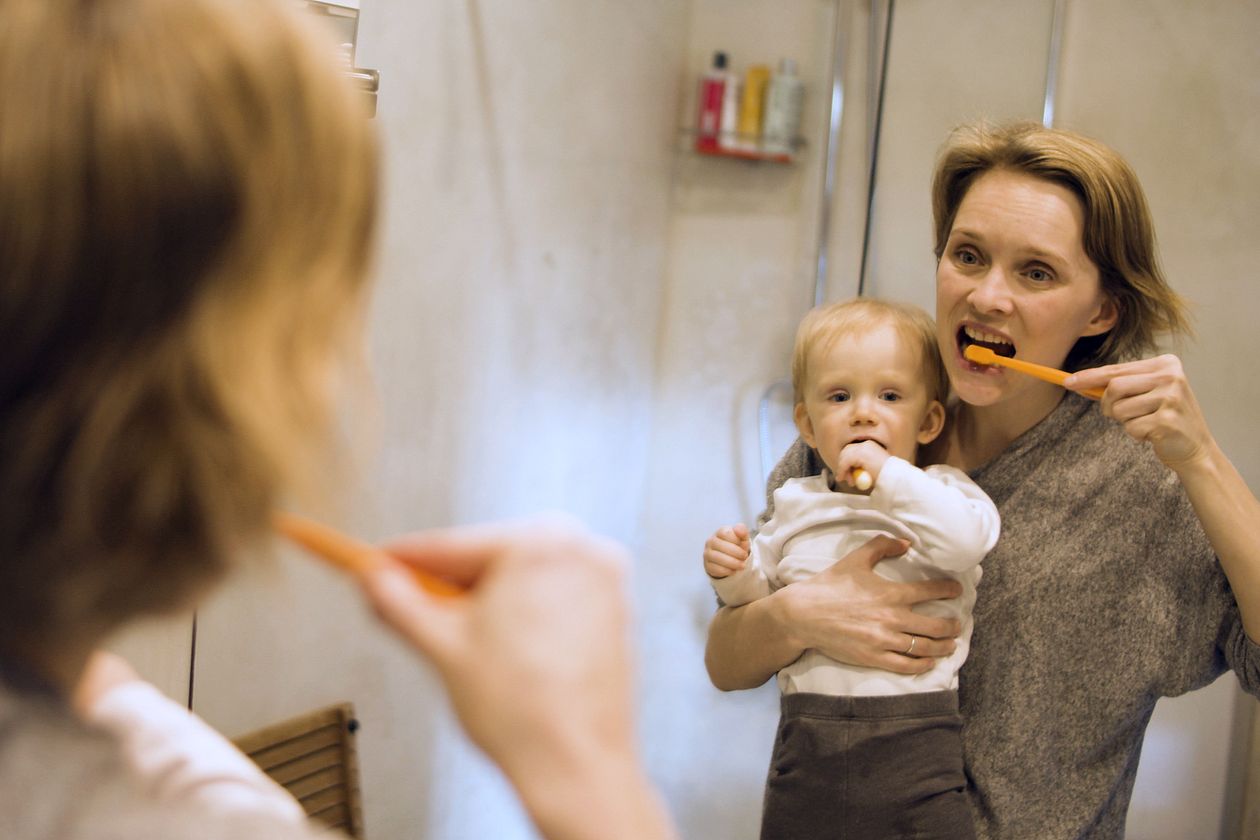 Mãe ajudando bebê a escovar os dentes no espelho do banheiro, contexto de melhor creme dental bebê