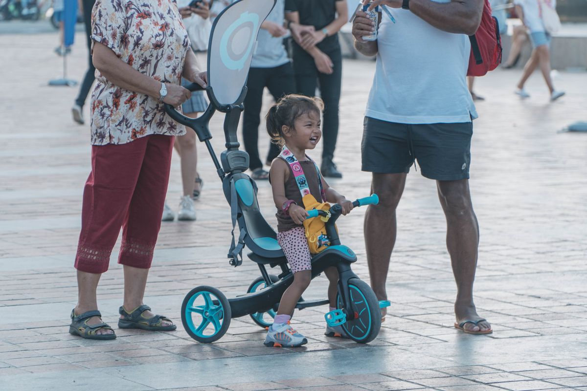 Criança em triciclo com a família na praça, contexto de uso do carrinho de passeio infantil ao ar livre
