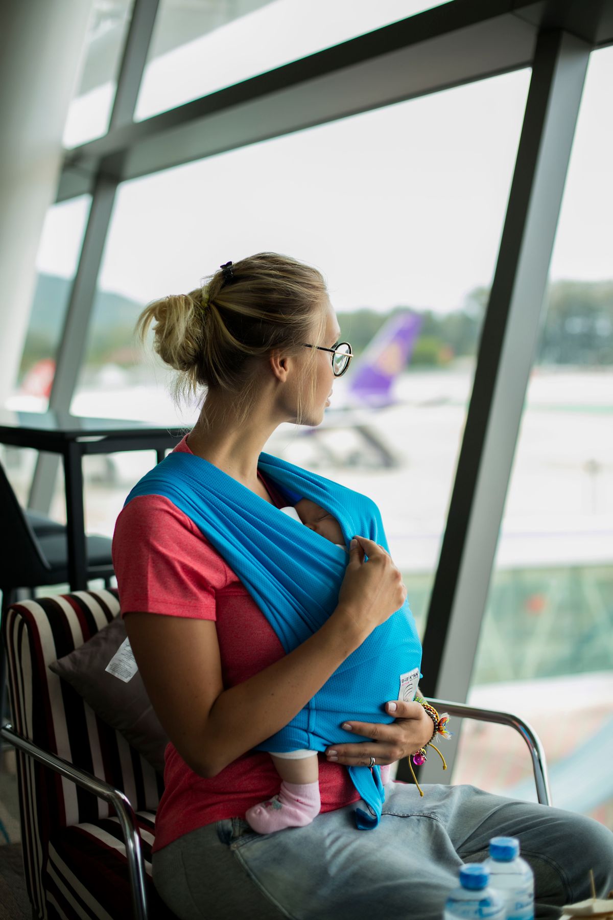 Mãe com bebê em canguru na janela do aeroporto, contexto de viagem de avião com bebê